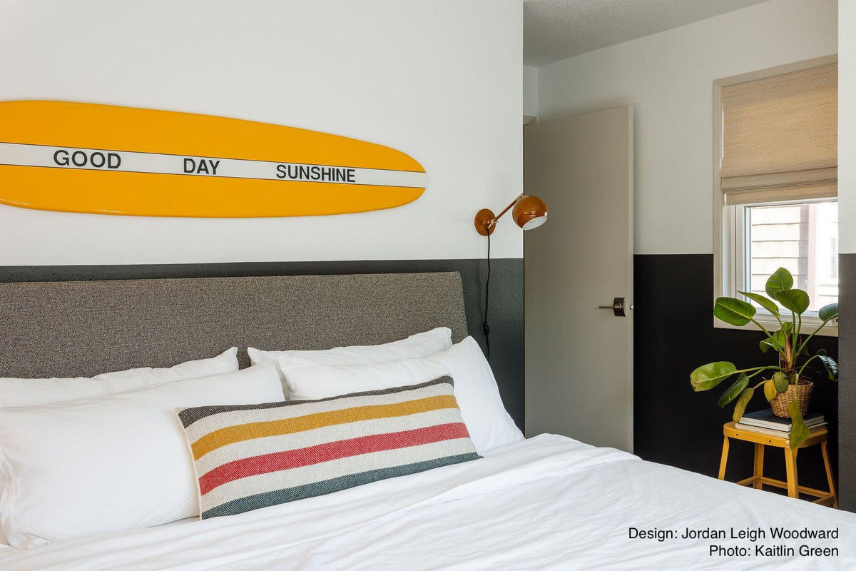 A bedroom with a bed that has white linens and a striped Glacier Park White pillow. A wall-mounted lamp and a small plant on a yellow stool are also visible.
