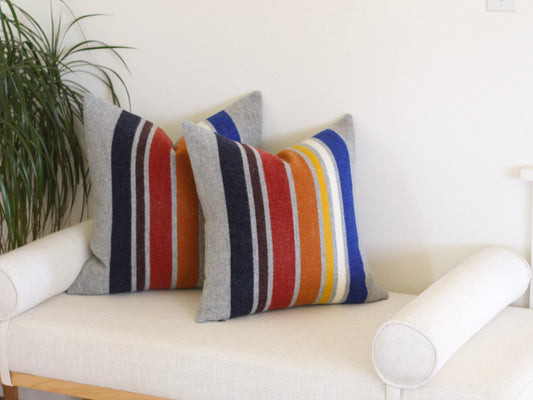Two pillow covers sitting on a white upholstered bench. The pillows feature colorful stripes on a gray background and are made with the Yosemite National Park pattern from Pendleton.