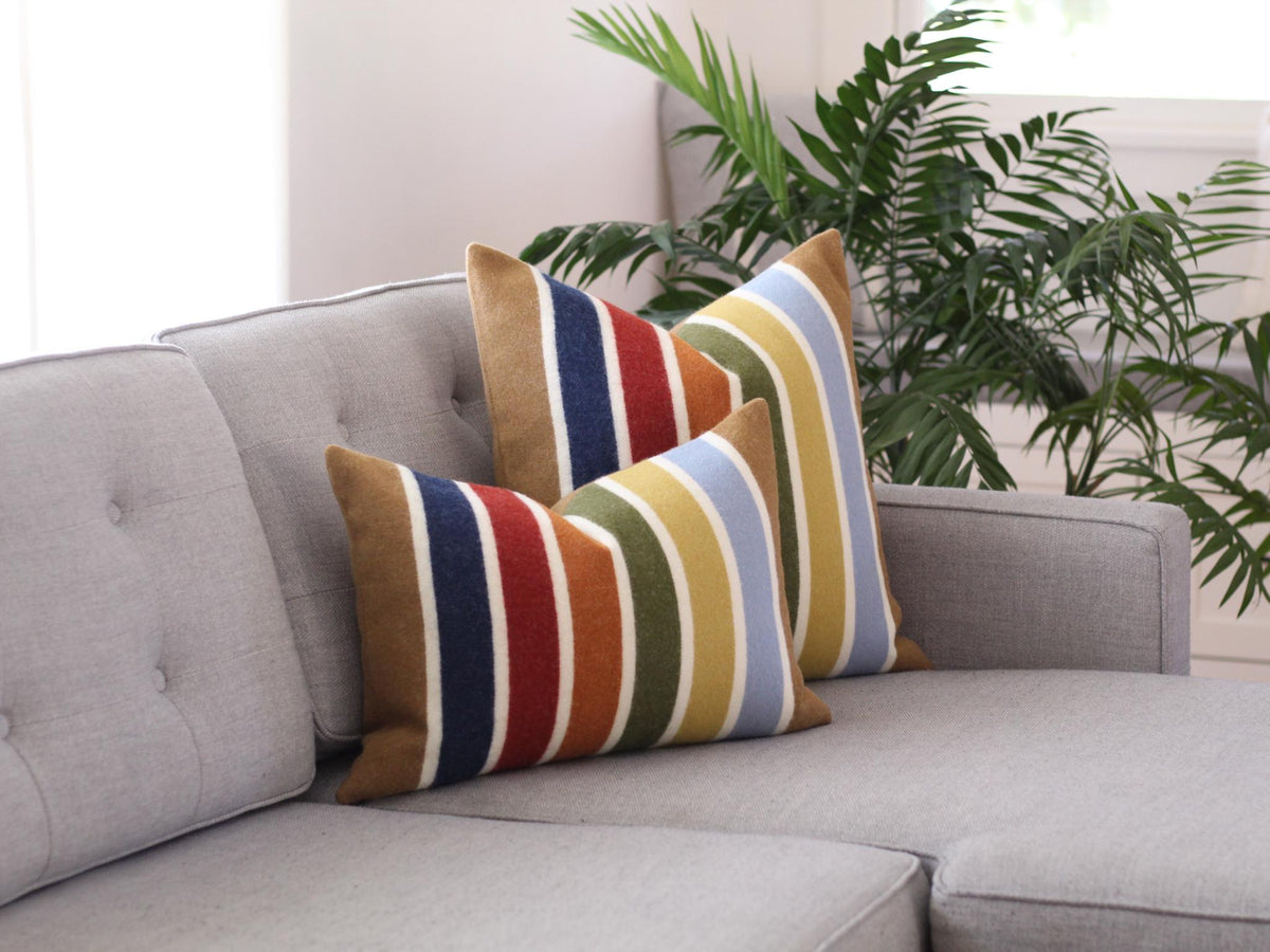 Two striped throw pillows on a gray couch. The pillows are made of wool and feature a multi-colored stripe pattern. The pillows have a camel brown, blue, red, orange, white, and green color scheme.