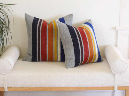Two decorative throw pillows with gray backgrounds and vertical stripes in navy, brown, red, orange, yellow, white, and blue. The pillows are placed on a white bench with cylindrical bolsters on each side.