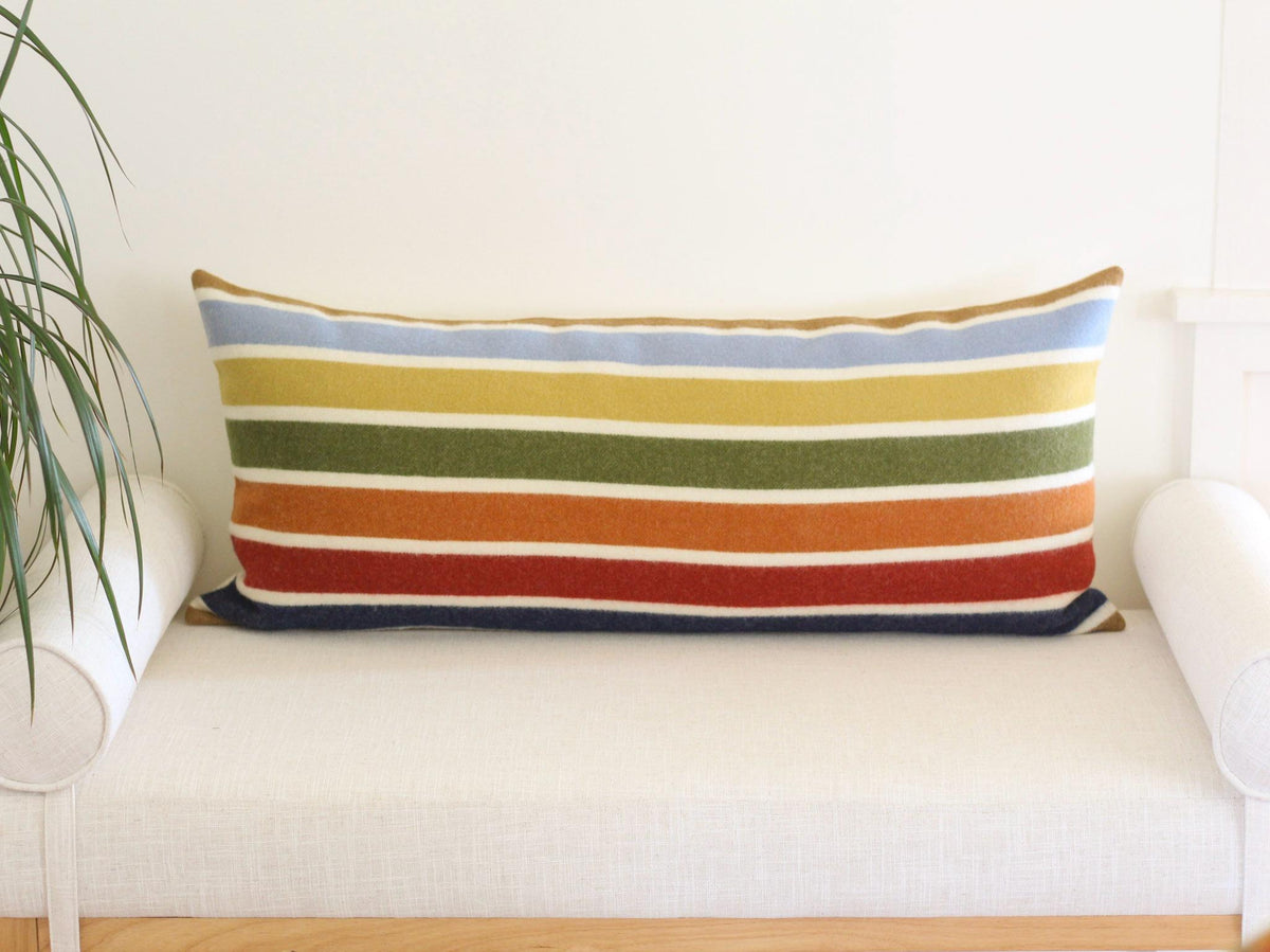 A rectangular pillow with horizontal stripes in various colors, including blue, yellow, green, orange, and red on a camel brown background. The pillow is placed on a white bench. The pillow is designed for decorative purposes.