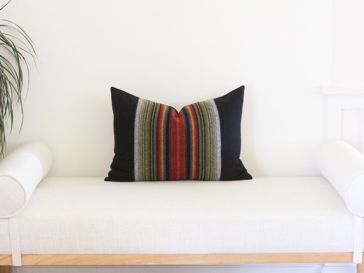 A rectangular throw pillow featuring solid black side panels and a central vertical stripe pattern. The stripes consist of thin, multi-colored lines in shades of red, orange, yellow, green, blue, and grey, creating a textured appearance.