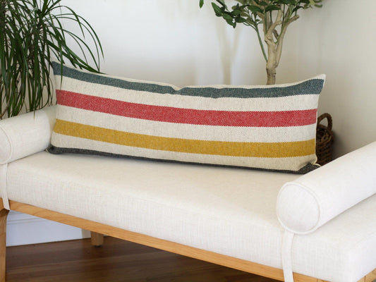 A rectangular lumbar pillow with horizontal stripes in red, yellow, green, and blackray. The pillow is placed on a white bench. The pillow's design adds a pop of color to the neutral-toned furniture.