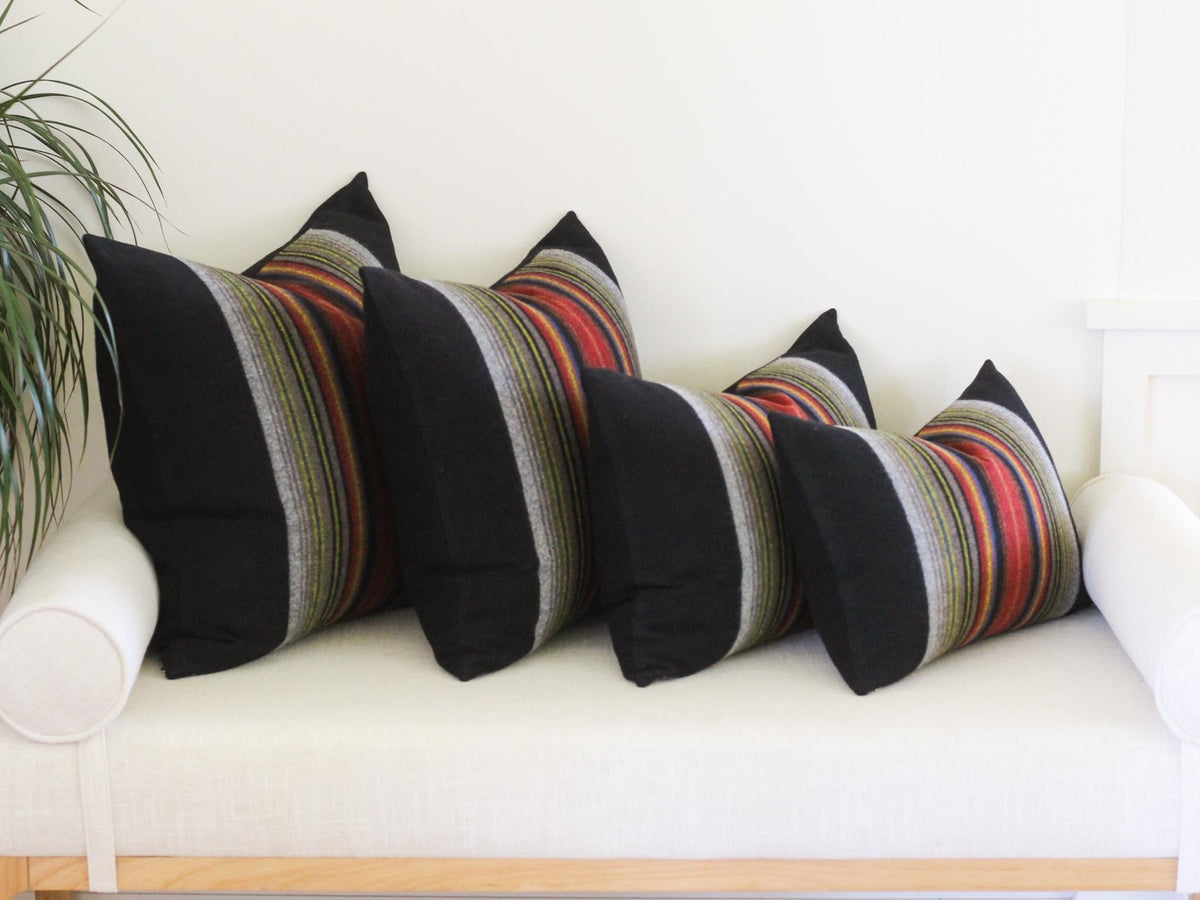 Four squre throw pillows arranged in a row, each featuring a solid black side and a vertical striped section in shades of red, orange, yellow, green, and grey. The pillows vary in size, decreasing from left to right in a line.