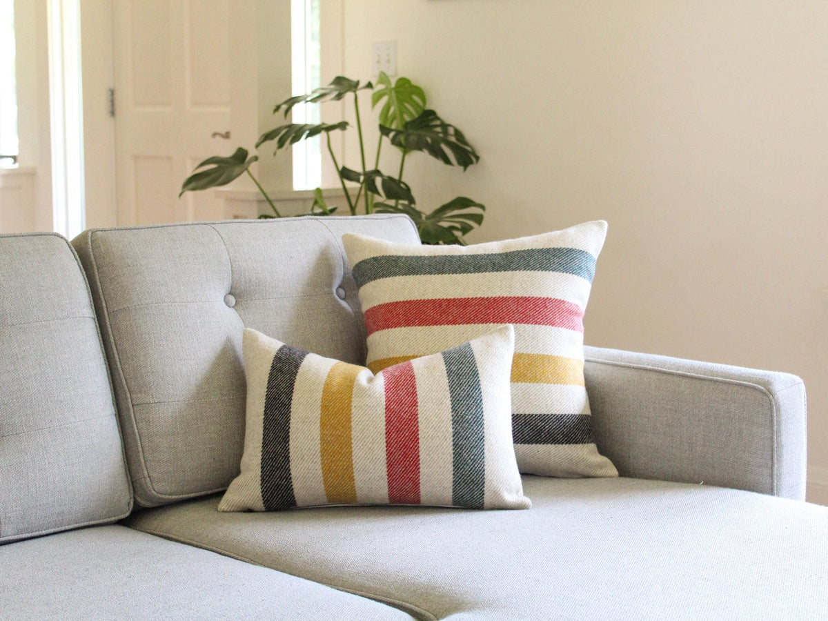 Two decorative throw pillows with a striped pattern in black, yellow, red, and green on a oatmeal and cream background. The pillows are placed on a gray sofa, creating a cozy and inviting atmosphere. The pillows add a pop of color and texture to the living space.