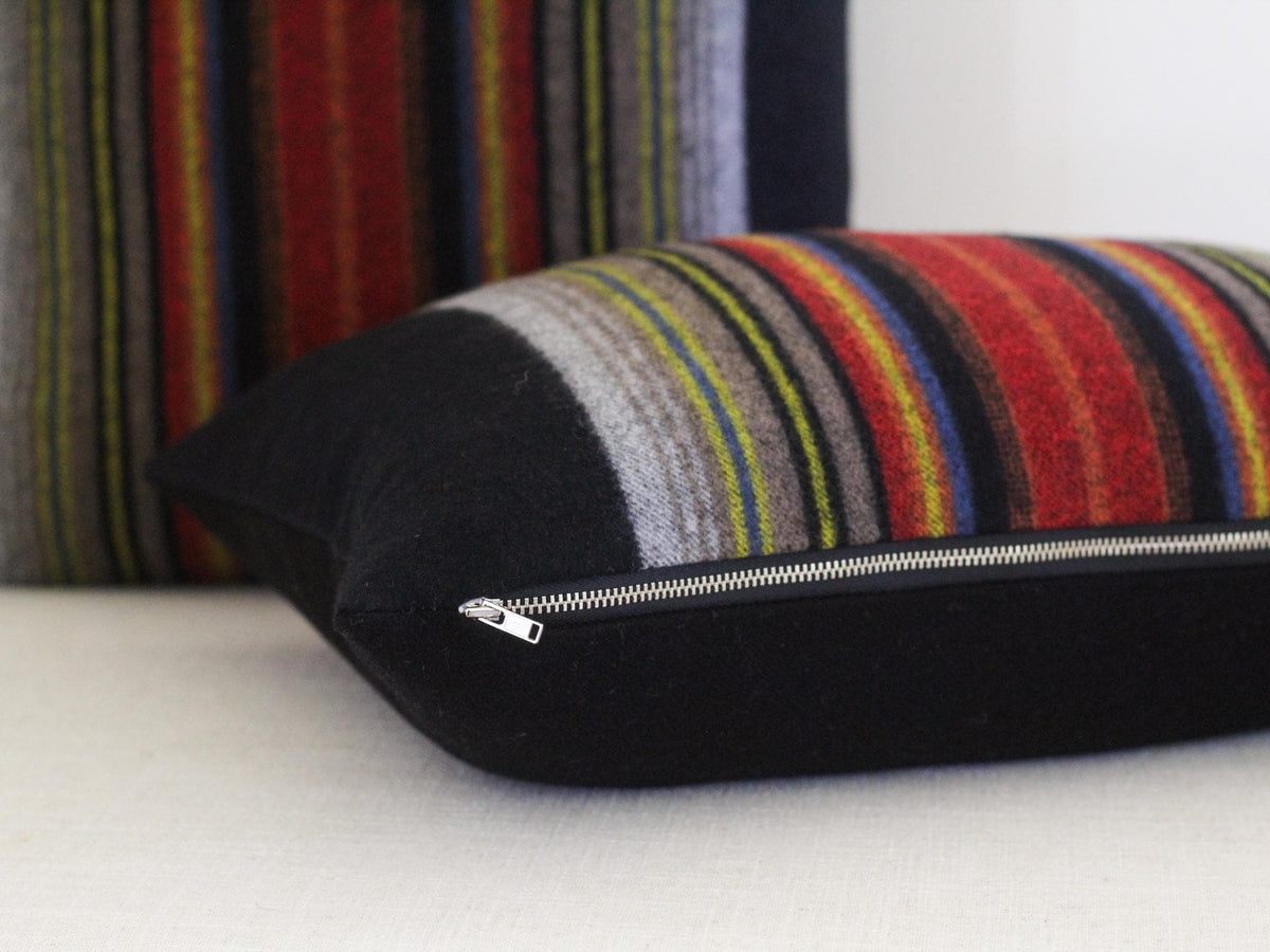 A rectangular throw pillow featuring a black section and a multi-colored striped section with red, yellow, blue, and grey tones. The pillow includes a visible silver metal zipper along the bottom seam. A second matching pillow is partially visible in the background.