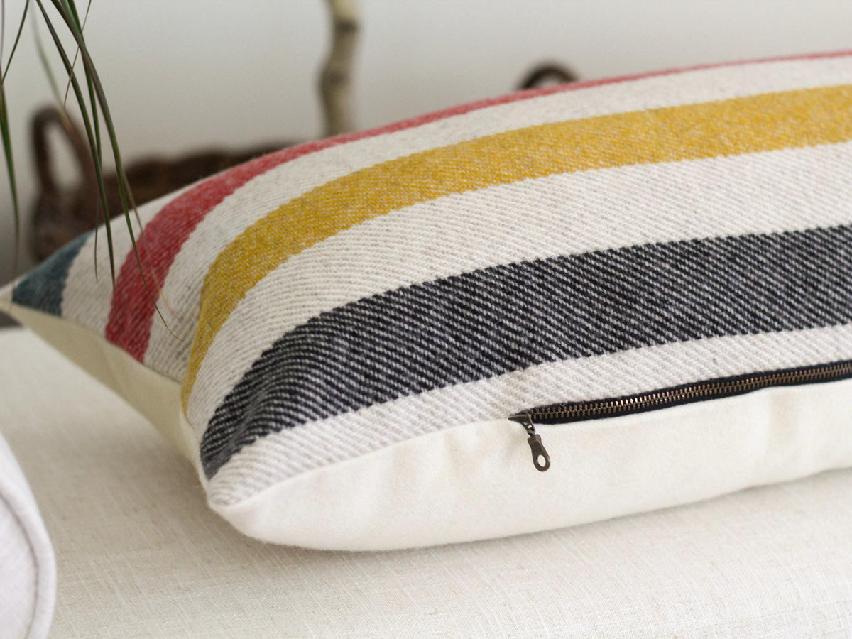 Rectangular pillow with a striped pattern in green, red, yellow, black. The pillow has a cream-colored base and a dark zipper closure along the bottom side.