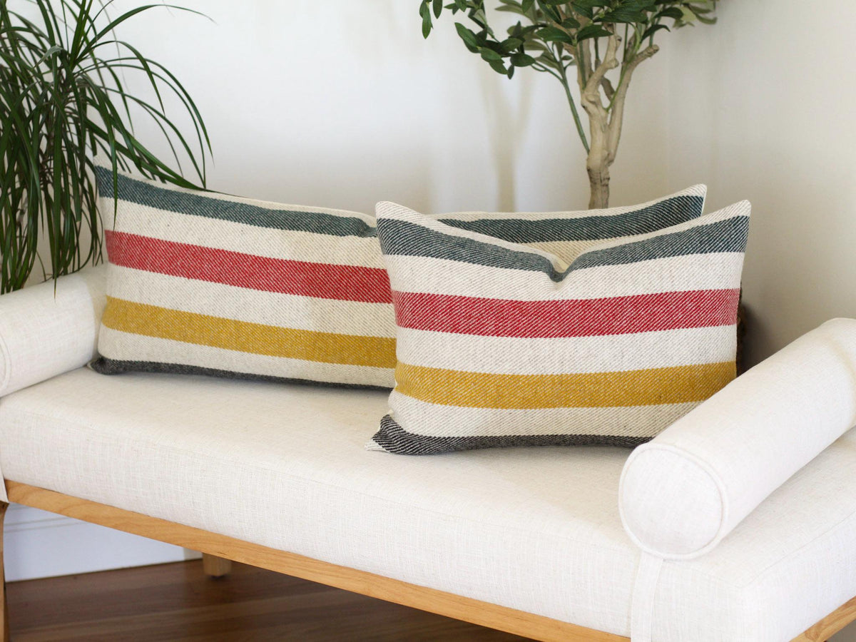 Two rectangular throw pillows with horizontal stripes in green, red, yellow, and black on a cream background. The pillows are on a white bench with a wooden frame.