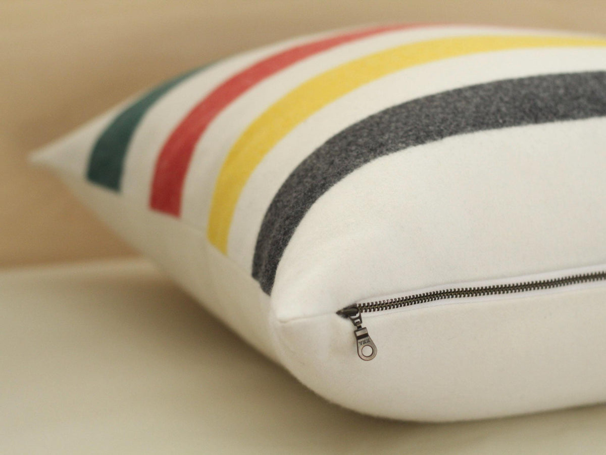 Cream decorative pillow with horizontal stripes in red, yellow, black, and green. The pillow has an exposed metal zipper closure and a soft, textured appearance. The pillow is designed for home decor.