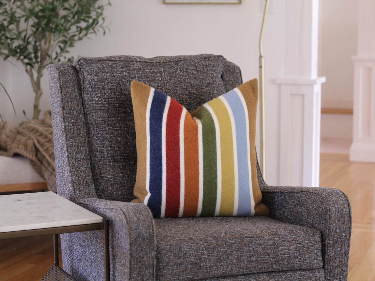 A gray armchair with a striped throw pillow featuring a rainbow of colors, including camel brown, blue, red, yellow, green, and brown. The pillow is placed on the chair's seat.
