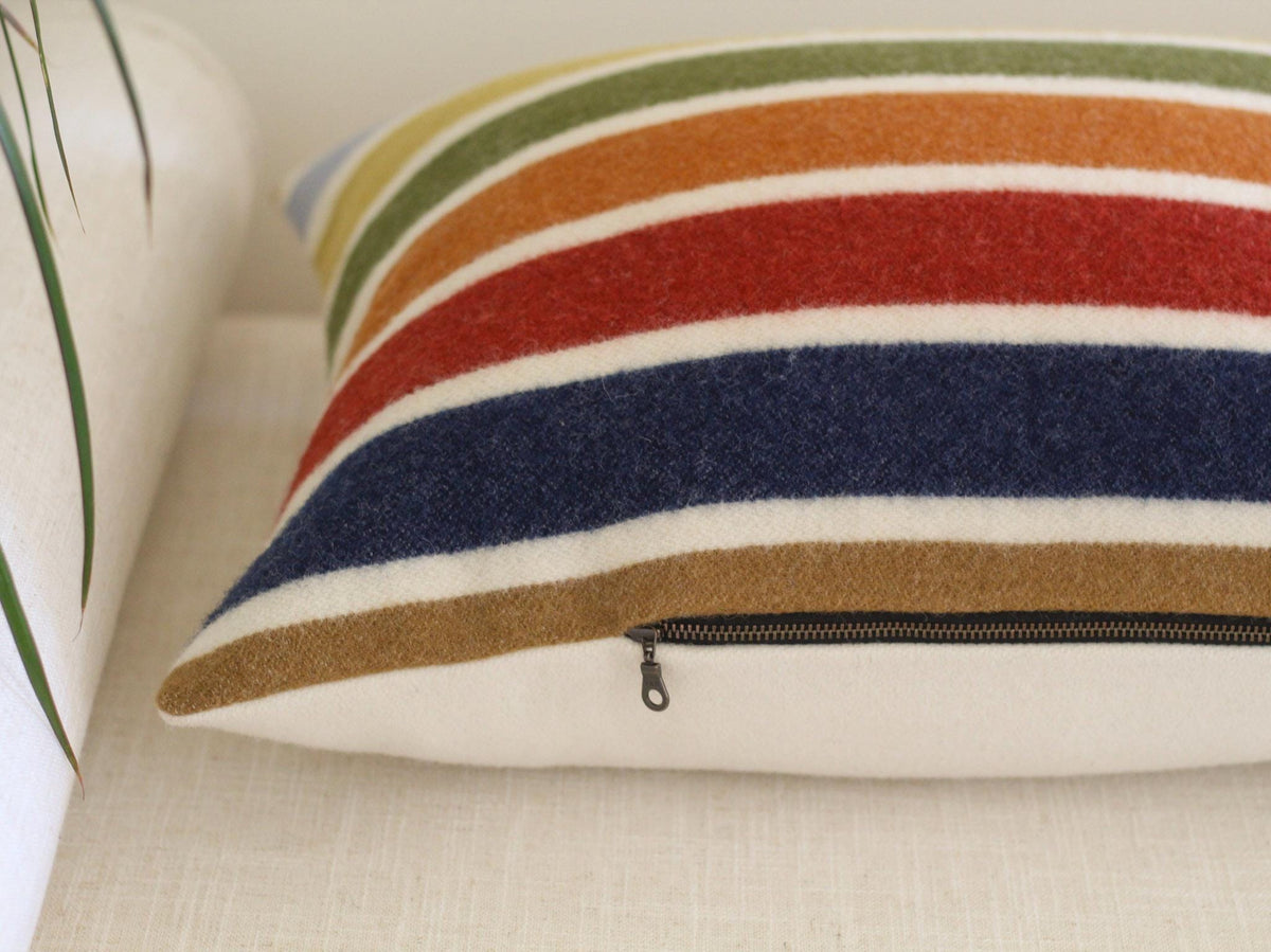 A decorative pillow with a striped pattern in shades of blue, red, orange, green, and camel brown. The pillow has a solid cream back and a exposed metal zipper closure. The pillow is a home decor item.