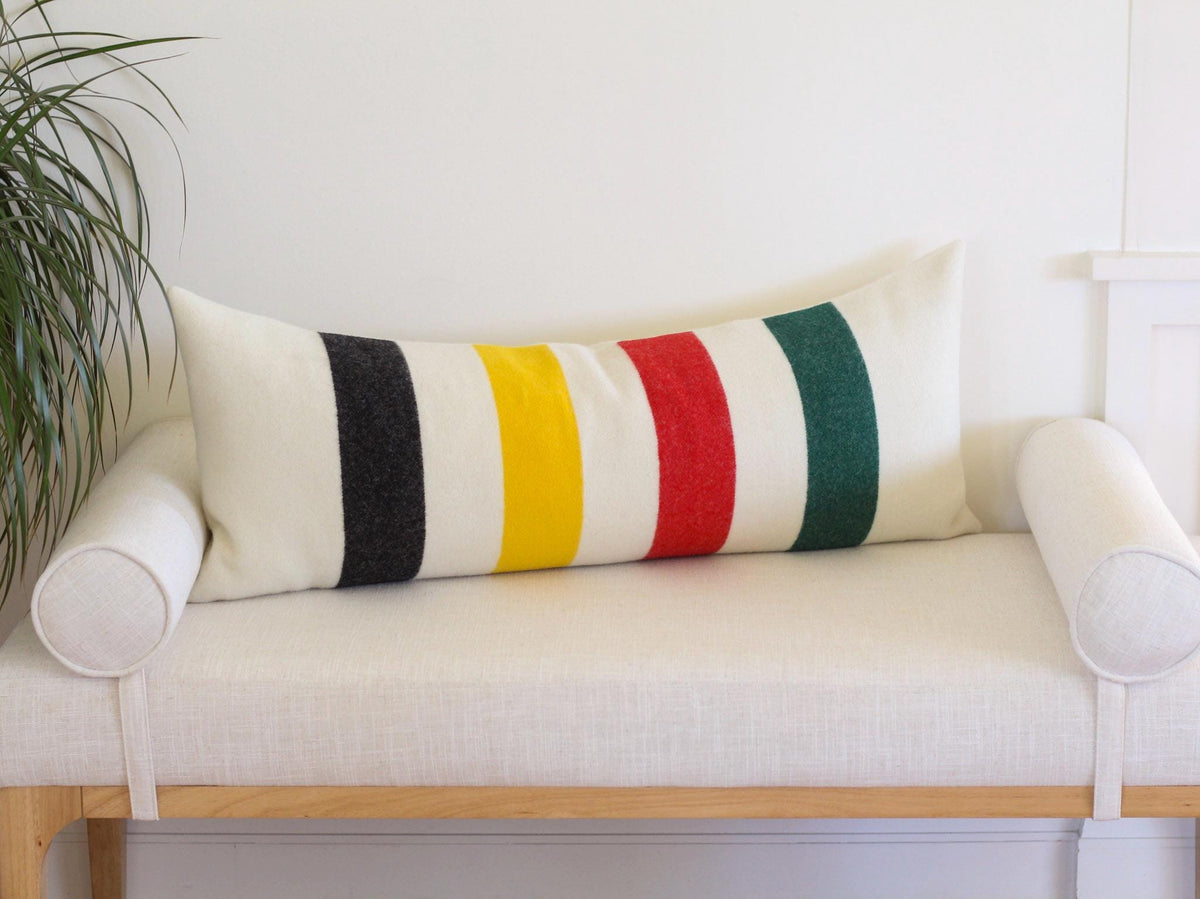 A rectangular lumbar pillow featuring a cream-colored base with vertical stripes in black, yellow, red, and green. The pillow is positioned on a light-colored bench with cylindrical bolster pillows on each side.