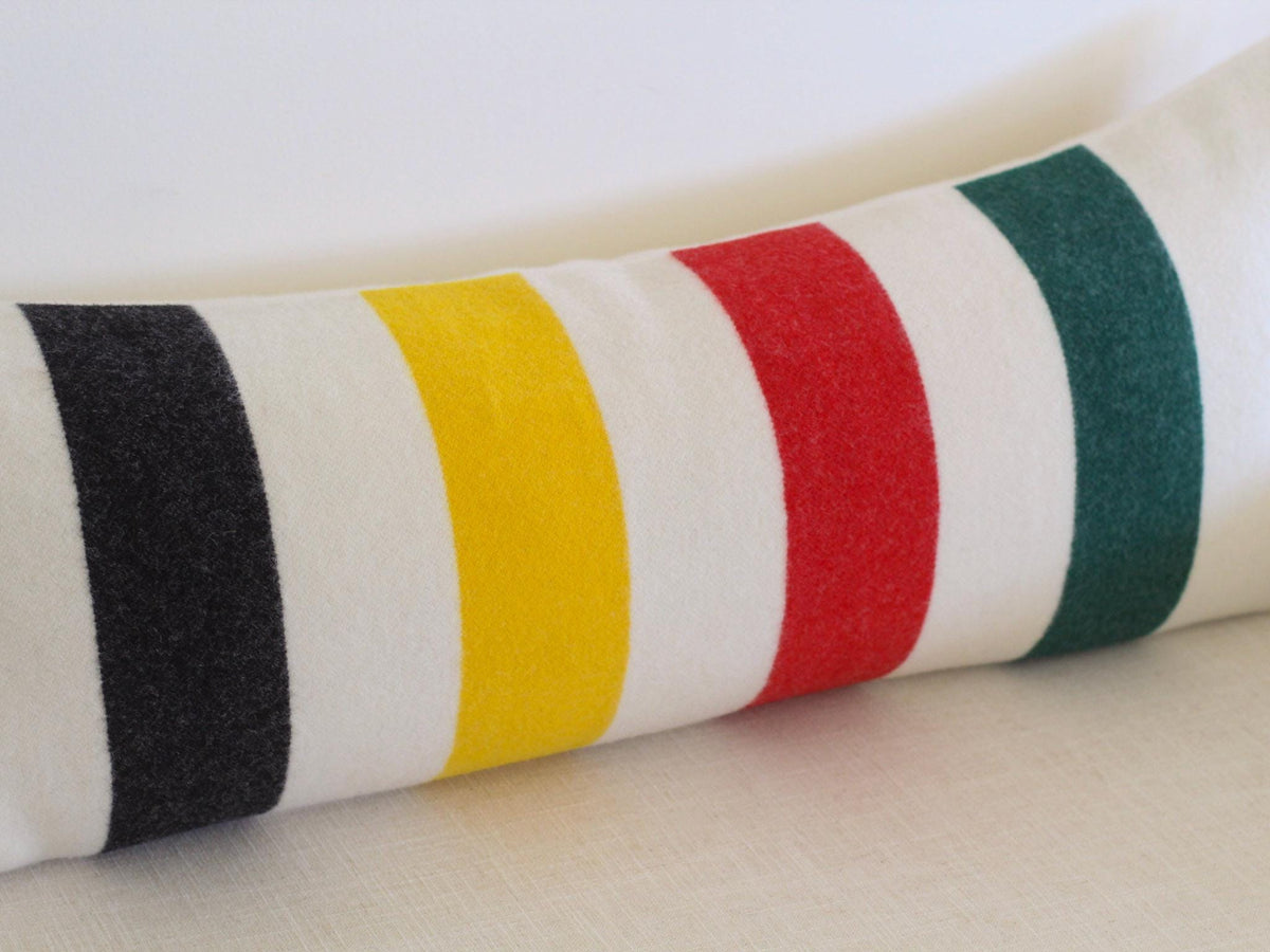 A long, rectangular bolster pillow featuring a cream-colored base with four distinct vertical stripes in black, yellow, red, and dark green.