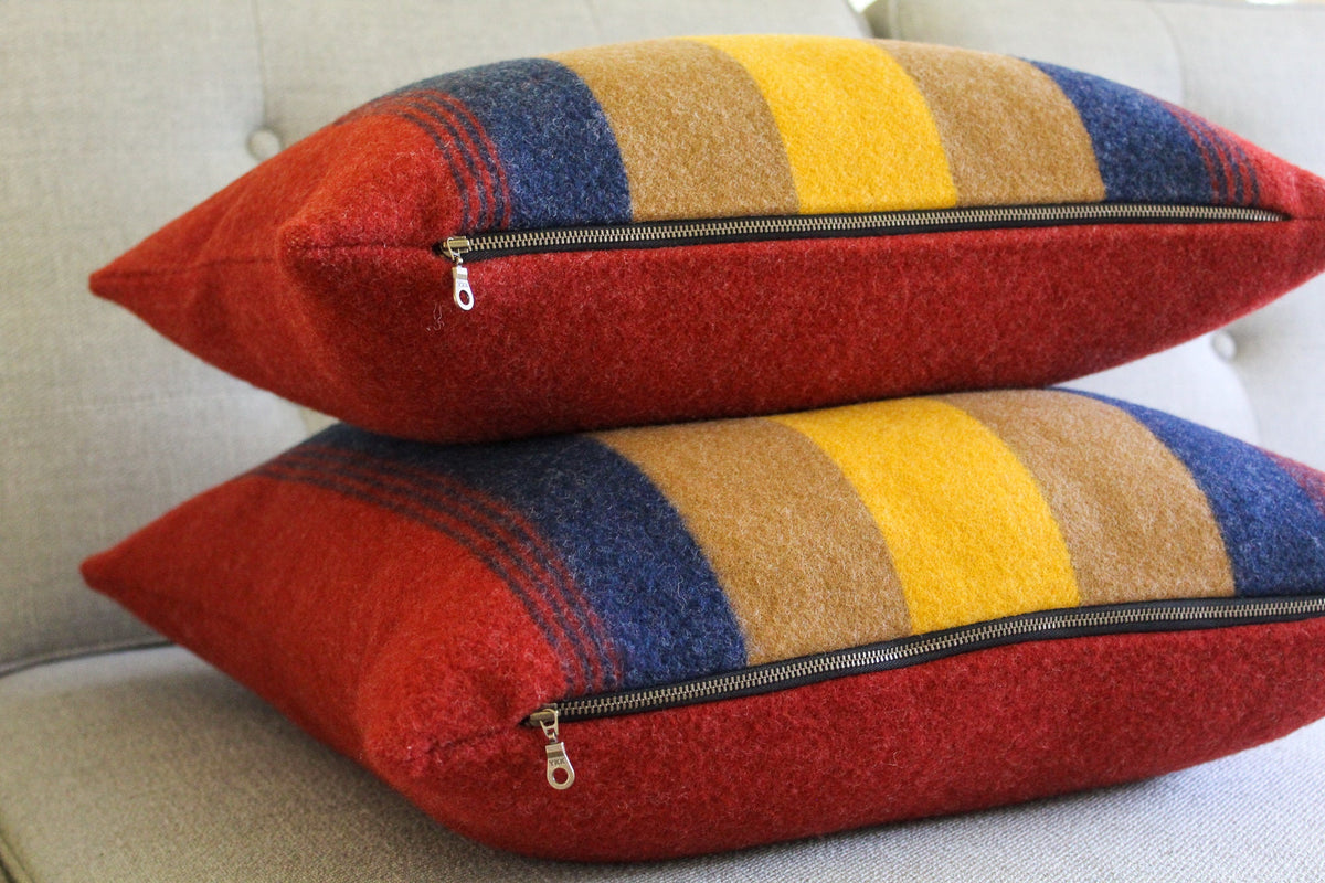 Two red, yellow, brown, and blue striped throw pillows with black zippers. The pillows are made of wool.
