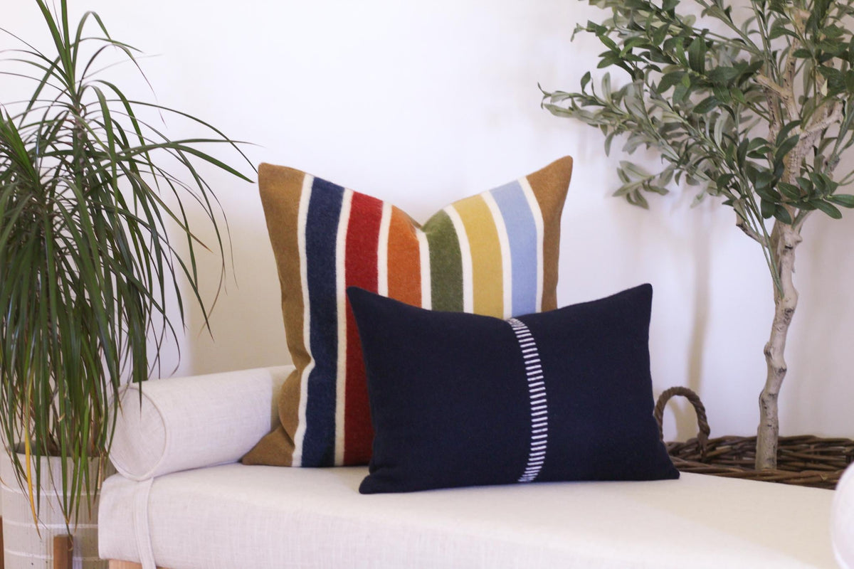 Two decorative throw pillows on a white bench. The larger pillow has a striped pattern in shades of camel brown, red, blue, green, and yellow. The smaller pillow is navy blue with a white stripe down the center.
