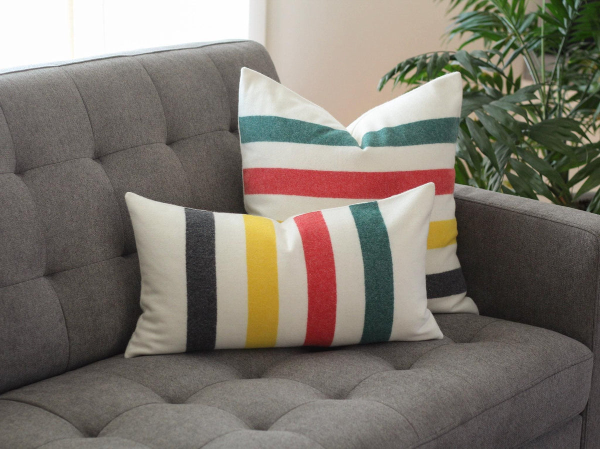 Two decorative pillows with a striped pattern in green, red, yellow, and black on a cream background. The pillows are placed on a gray tufted sofa. The pillows add a pop of color to the neutral-toned living room.