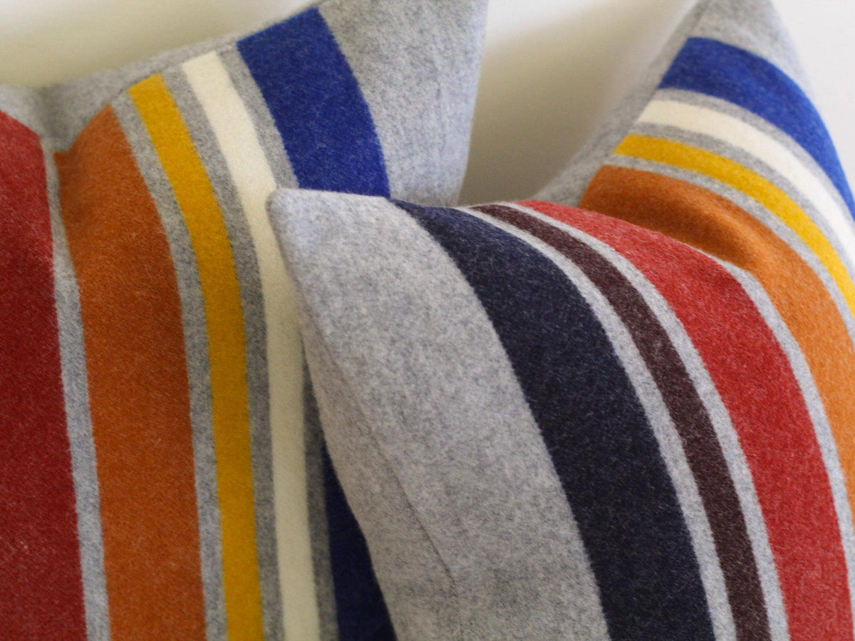 Close up of two gray throw pillows with vertical stripes in navy, brown, red, orange, yellow, white, and blue.  The pillows have a soft, textured appearance, suggesting a cozy, comfortable design.