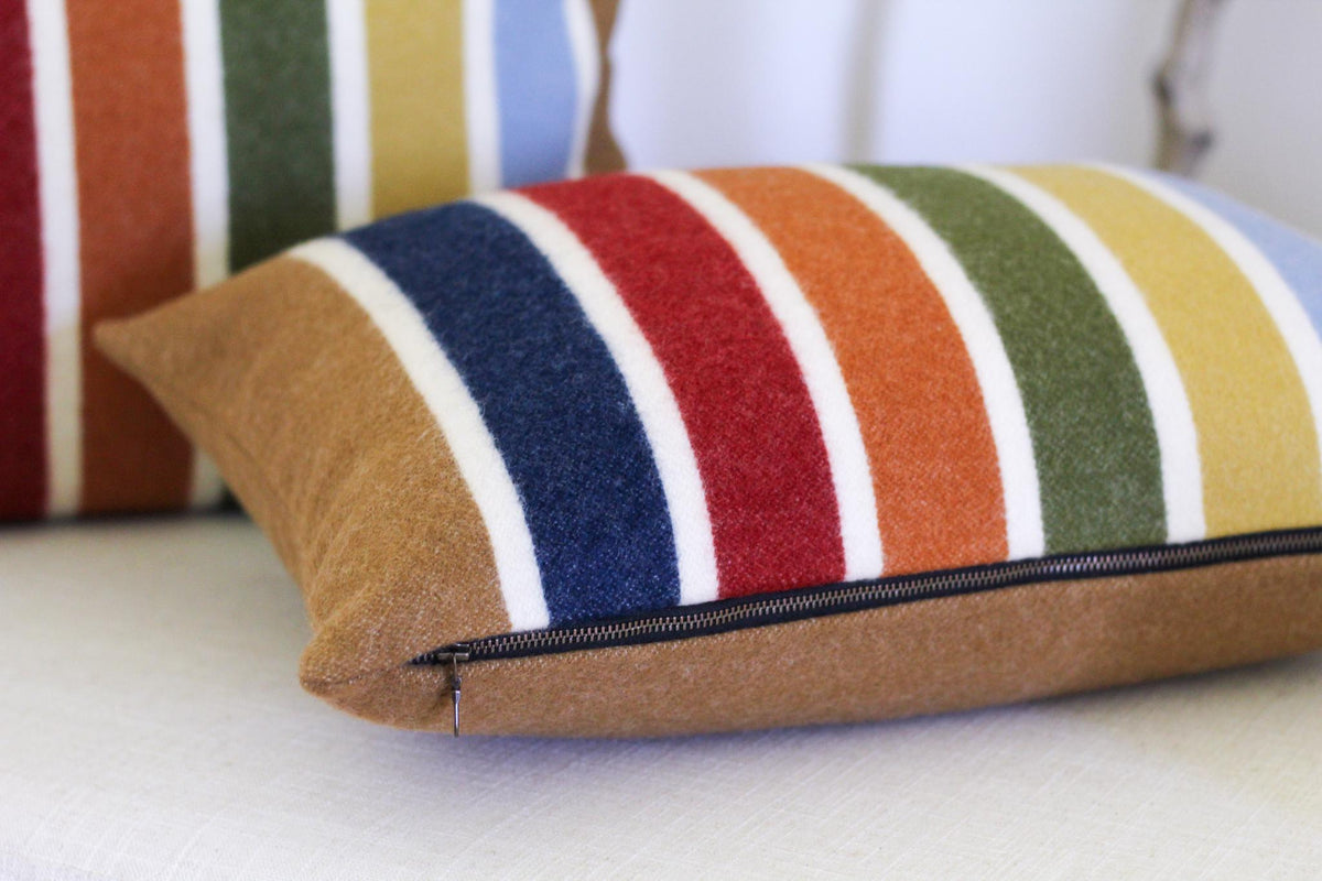 A brown striped wool throw pillow with a zipper closure. The pillow has stripes of white, red, orange, yellow, green, blue, and brown.