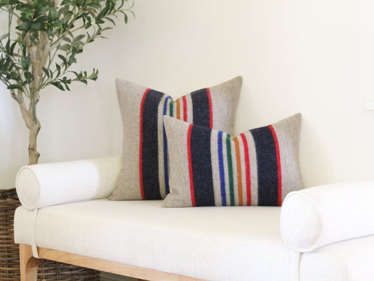 Two striped throw pillows with a beige background and navy blue, red, orange, green, and white stripes. The pillows are on a white bench with two white cylindrical pillows.