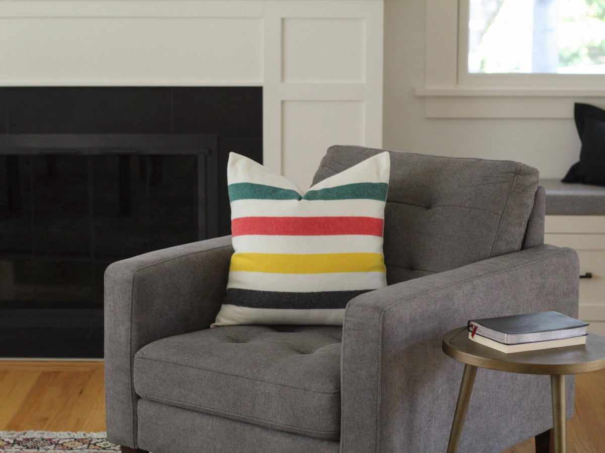A gray armchair with a striped pillow featuring green, red, yellow, and black stripes. A small, round, gold side table holds a book. The background includes a fireplace and a window.