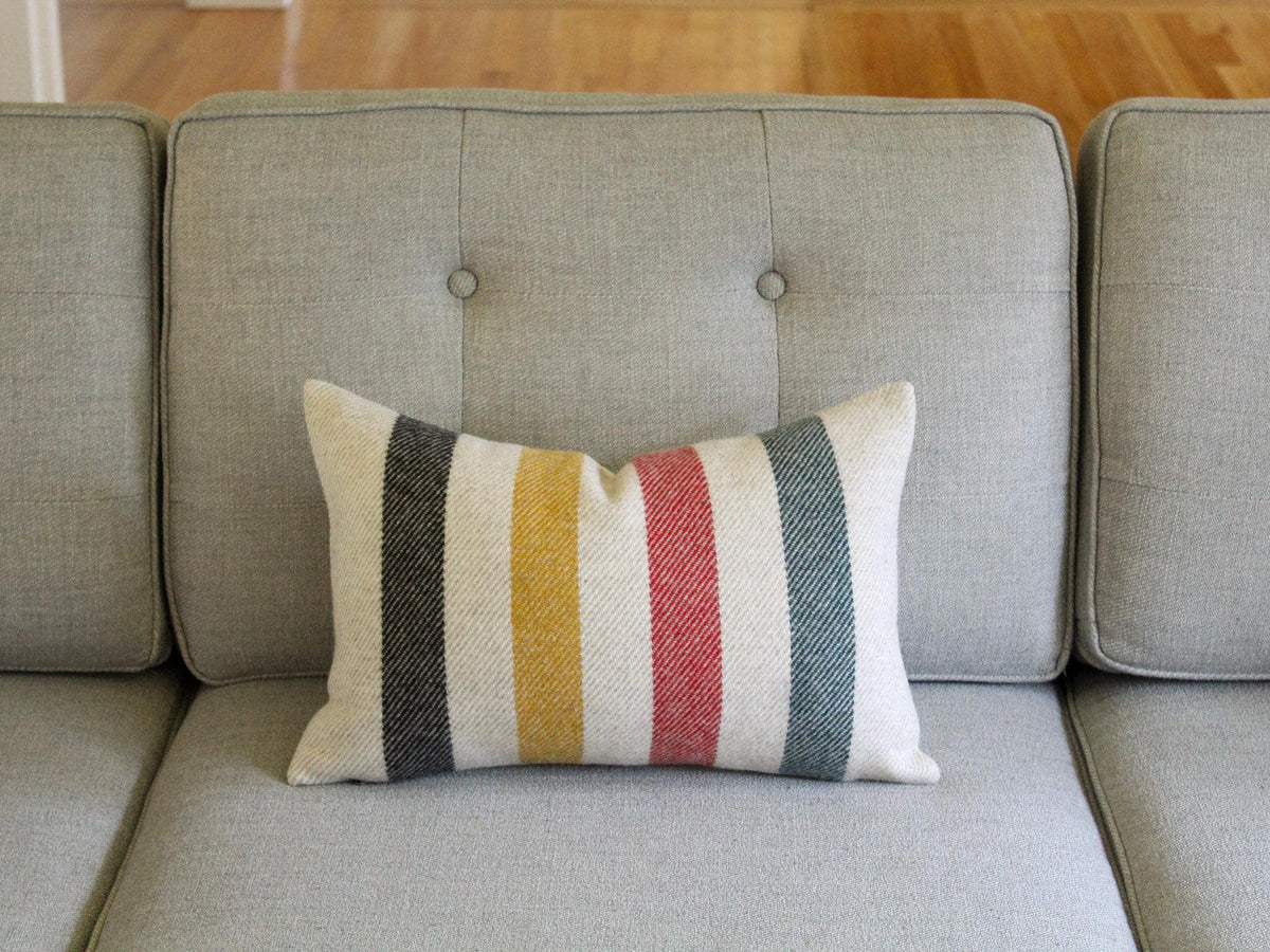 A rectangular decorative pillow with vertical stripes in black, yellow, red, and green on an oatmeal cream background. The pillow is placed on a gray sofa with button tufting.