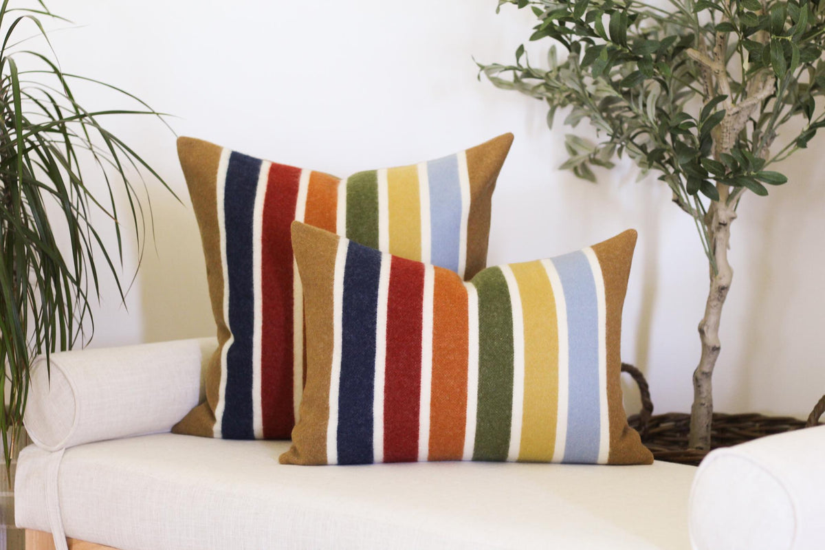 Two striped throw pillows with a variety of colors, including camel brown, blue, red, orange, yellow, and green. The pillows are on a white bench.