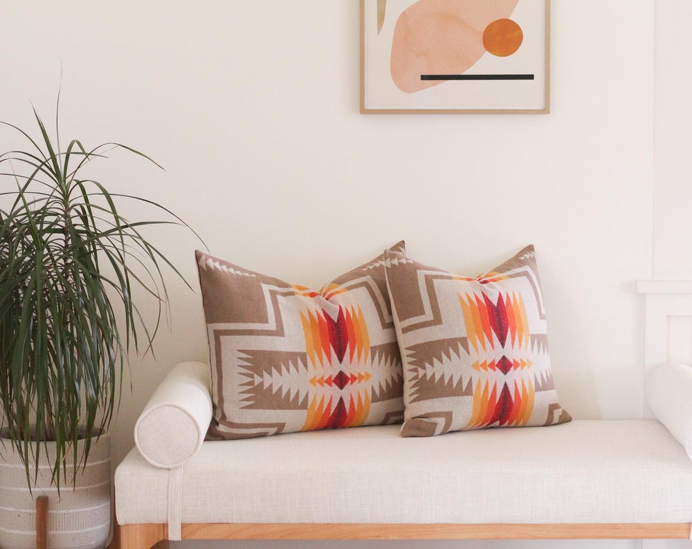 Two True Having pillow covers in the Harding Tan pattern, which features a native American motif in tan, cream, red, orange, and yellow. The pillow covers sit on a cream bench next to a large potted plant. Artwork hangs above the bench.