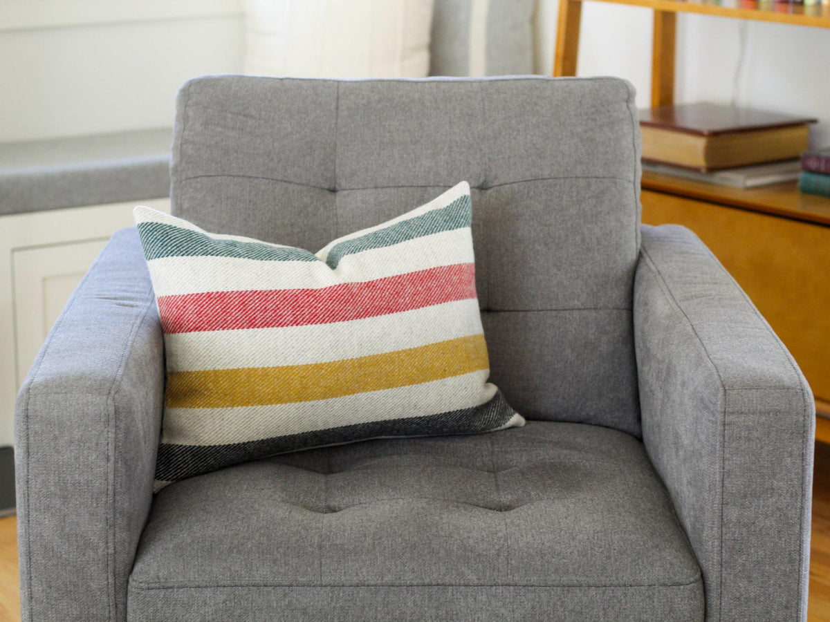 A gray upholstered armchair with a rectangular pillow. The pillow has horizontal stripes in green, red, yellow, and black with an oatmeal cream background. The chair has a button-tufted back and armrests.