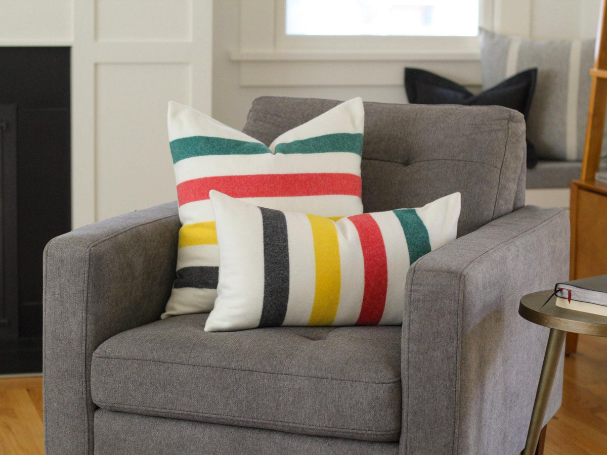 Two decorative throw pillows with a striped pattern in red, green, yellow, and black on a cream background. The pillows are placed on a gray armchair. The pillows add a pop of color and pattern to the neutral-toned room.