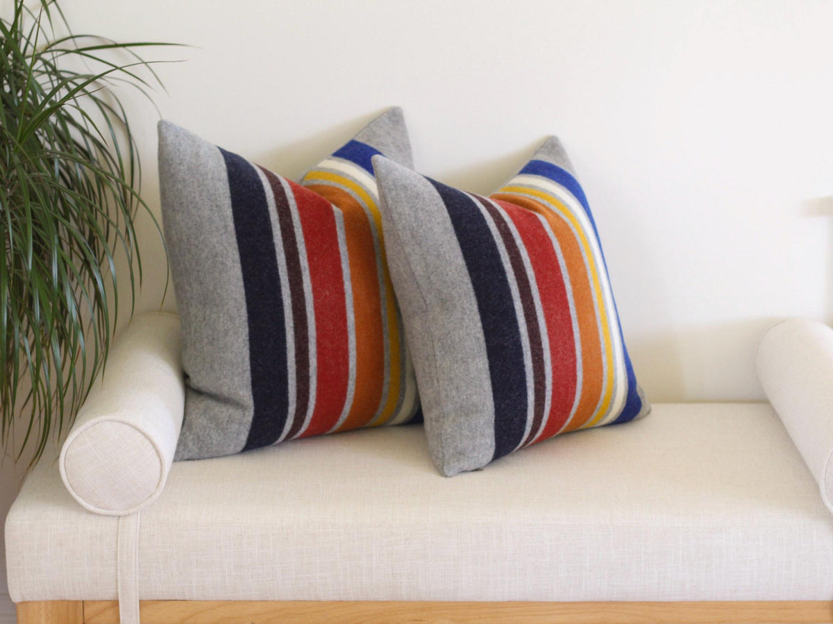 Two gray throw pillows with vertical stripes in navy, brown, red, orange, yellow, white, and blue. The pillows are on a white bench with a wooden frame. A green plant is visible on the left.
