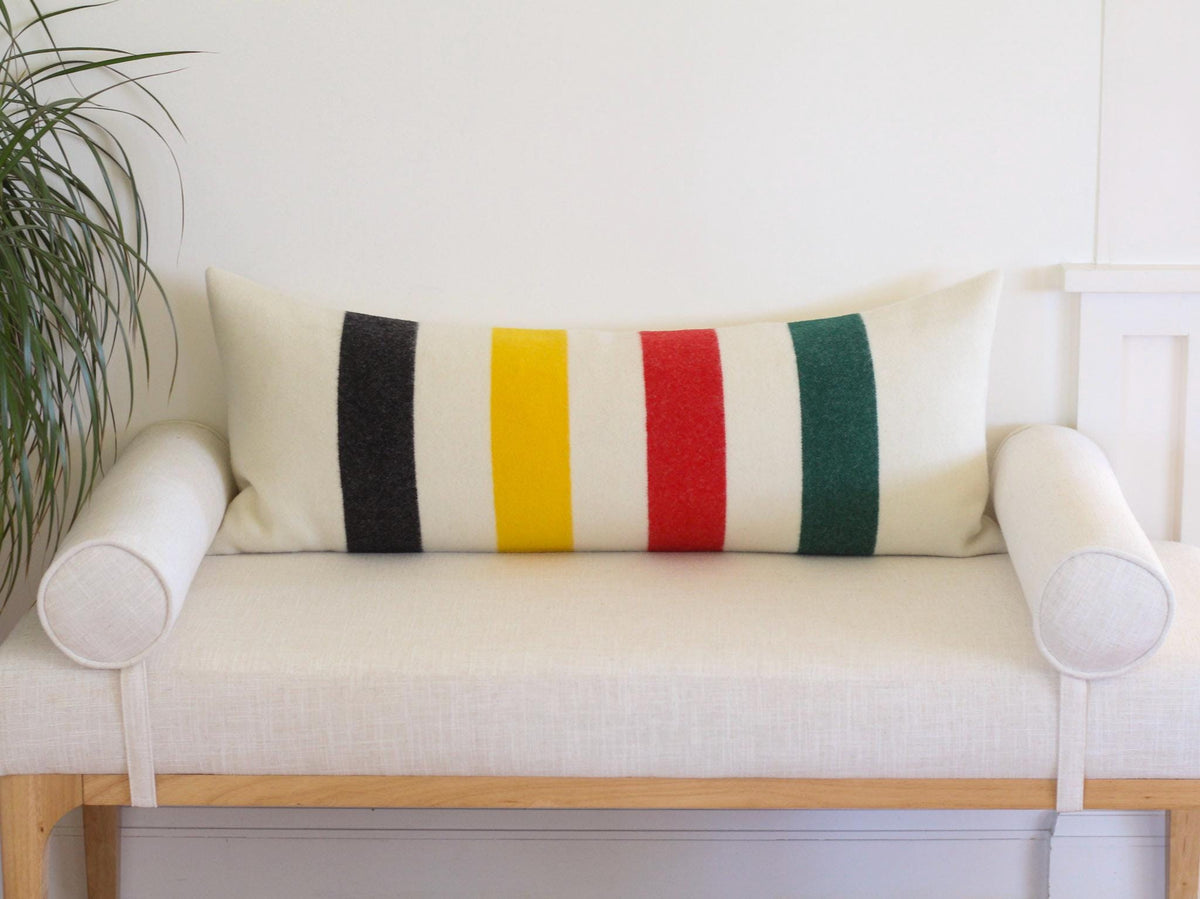 A rectangular lumbar pillow featuring a cream-colored base with vertical stripes in black, yellow, red, and green. The pillow is positioned on a light-colored bench with cylindrical bolster pillows on each side.