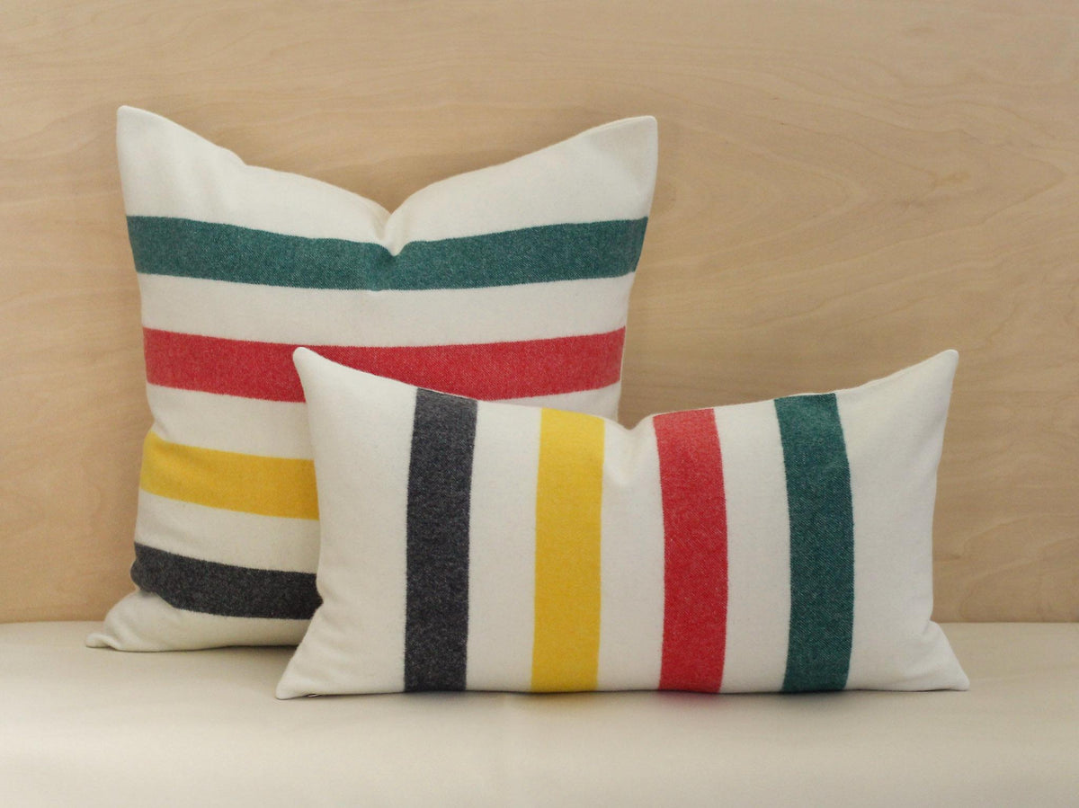Two decorative pillows with a cream-colored background and bold stripes in black, yellow, red, and green. One pillow is square, and the other is rectangular. 