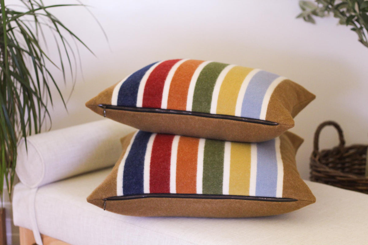 Two striped wool throw pillows with a stripe front and camel brown backing. The pillows are stacked on top of each other. The pillows have camel brown, blue, red, orange, green, yellow, and white stripes.
