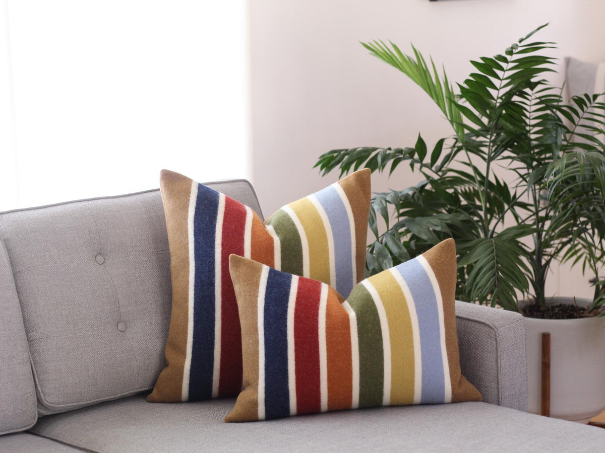 Two striped throw pillows with a colorful pattern of camel brown, blue, red, yellow, green, and white stripes on a gray couch.