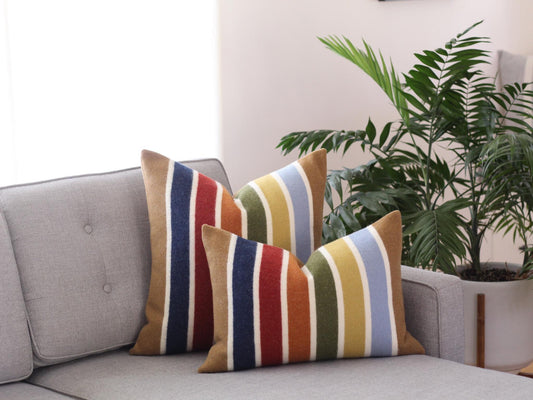 Two striped throw pillows with a colorful pattern of camel brown, blue, red, yellow, green, and white stripes on a gray couch.