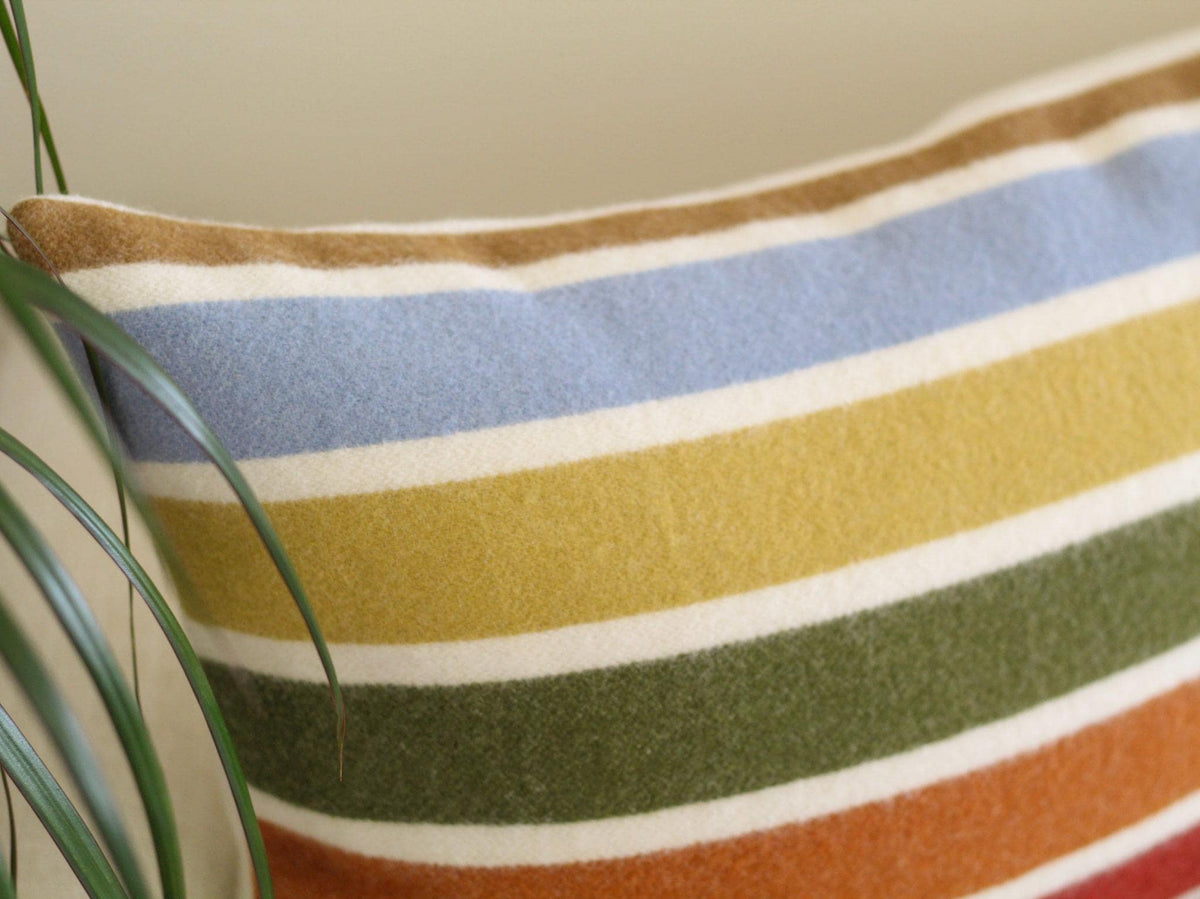 Close-up of a rectangular pillow with horizontal stripes in various colors. The stripes include white, light blue, yellow, green, and orange. The pillow has a camel brown border and is made of a soft, textured fabric.