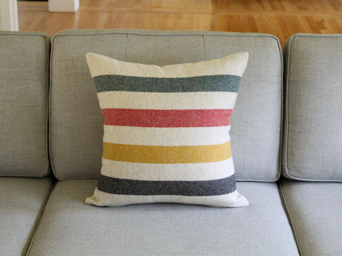 A square decorative pillow with horizontal stripes in various colors, including green, red, yellow, and black, on an oatmeal cream background. The pillow is placed on a gray sofa, adding a pop of color to the neutral setting.