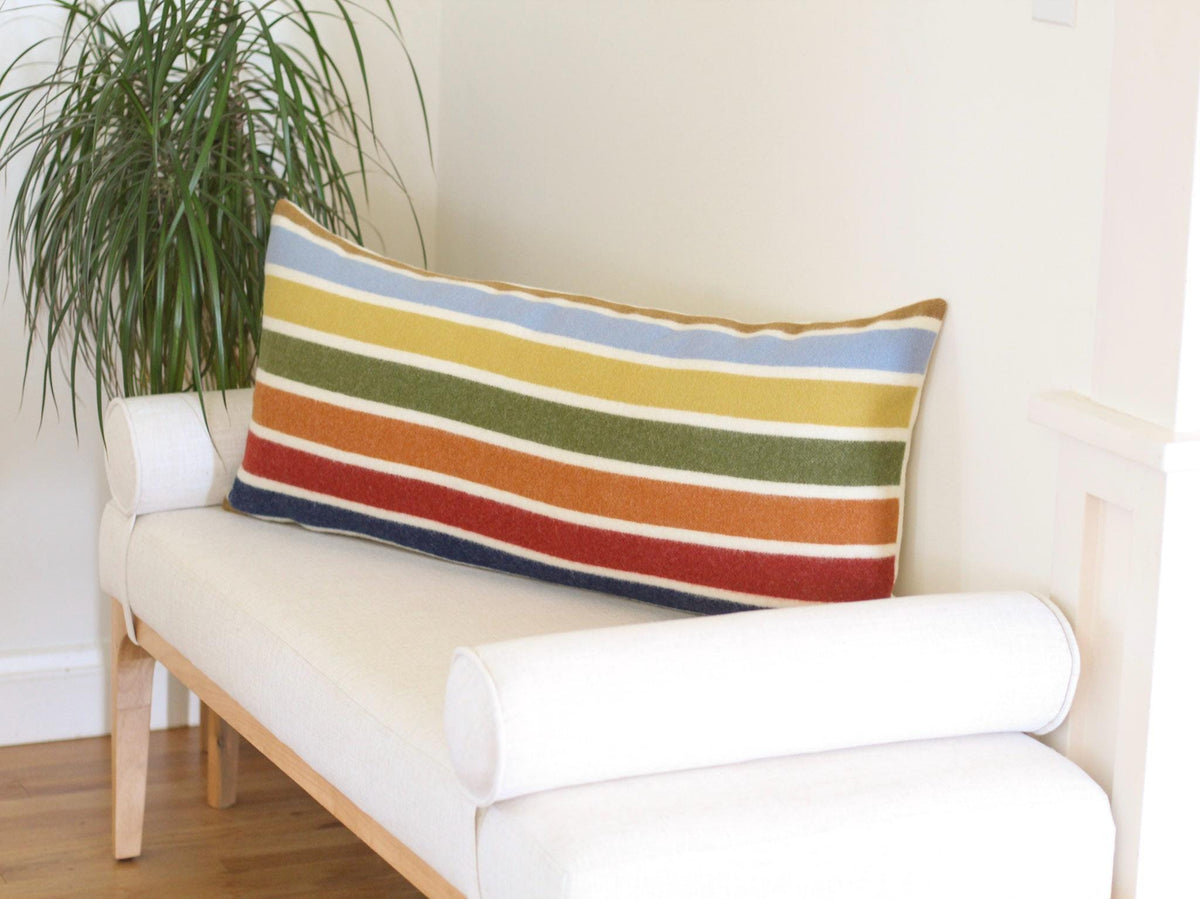 A rectangular pillow with horizontal stripes in various colors, including camel brown, blue, yellow, green, red, and orange. The pillow is placed on a white bench with rounded bolster pillows on each end.