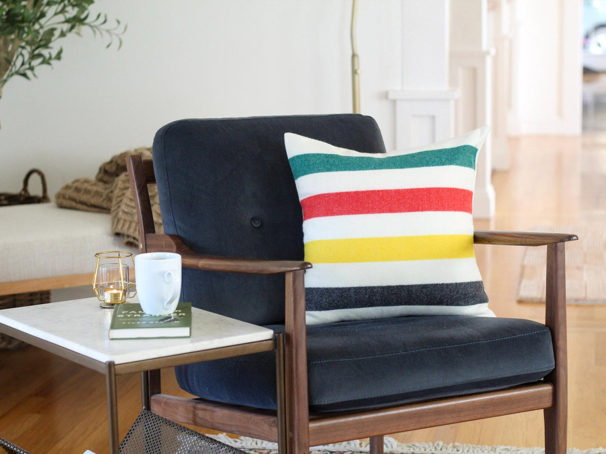 A navy blue armchair with wooden arms and a colorful striped pillow. The pillow features horizontal stripes of green, red, yellow, and black. A small side table holds a book, a cup, and a candle.
