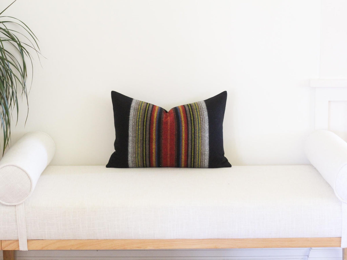 A rectangular throw pillow featuring solid black side panels and a central vertical stripe pattern. The stripes consist of thin, multi-colored lines in shades of red, orange, yellow, green, blue, and grey, creating a textured appearance.