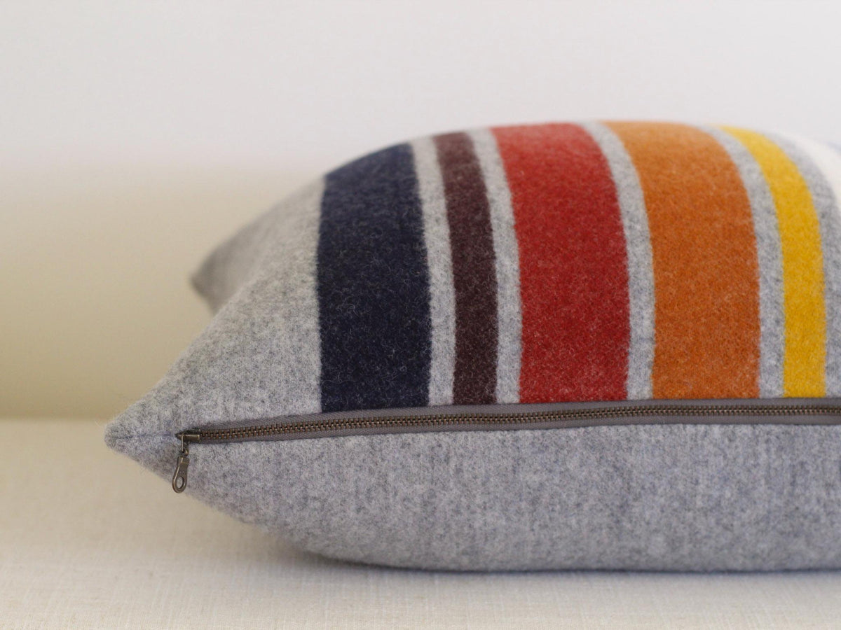 A square pillow with a gray base and colorful vertical stripes. The stripes include navy, brown, red, orange, yellow, white, and blue. A zipper runs along the bottom edge of the pillow.