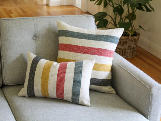 Two decorative throw pillows with a striped pattern in black, yellow, red, and green on a oatmeal and cream background. The pillows are placed on a gray sofa, creating a cozy and inviting atmosphere. The pillows add a pop of color and texture to the living space.