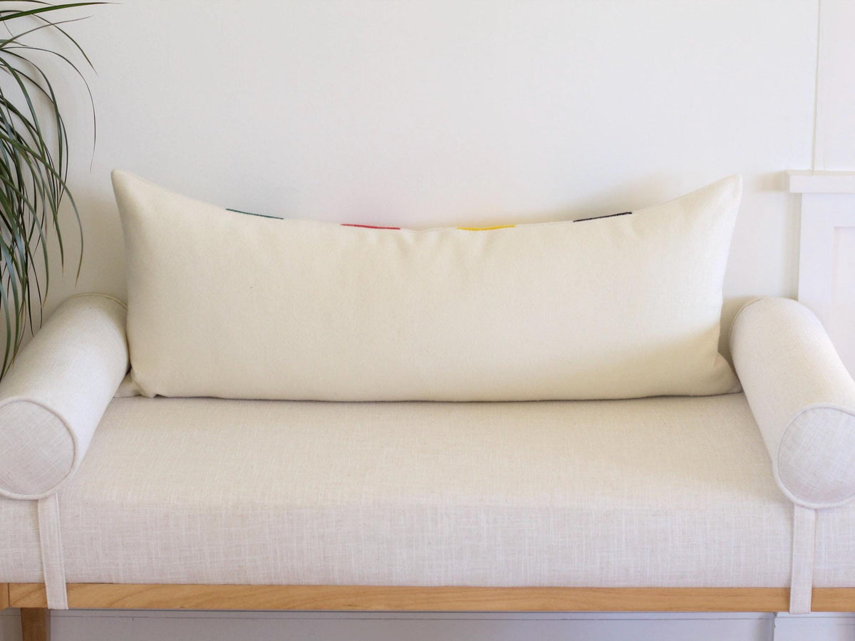 The back side of a rectangular lumbar pillow in solid cream. The pillow is positioned on a light-colored bench with cylindrical bolster pillows on each side.