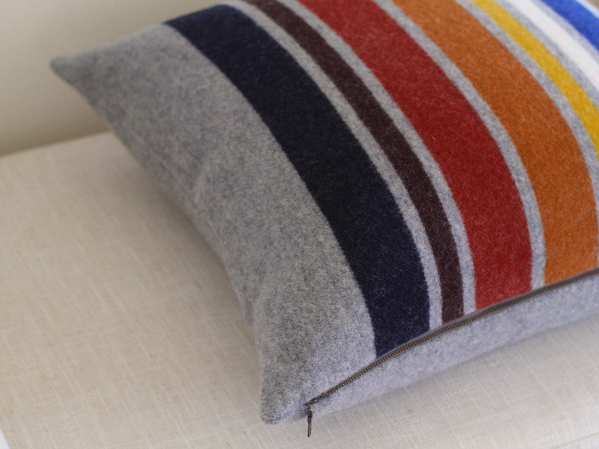 A square pillow with a gray base and colorful vertical stripes. The stripes include navy, brown, red, orange, yellow, white, and blue. A zipper runs along the bottom edge of the pillow.