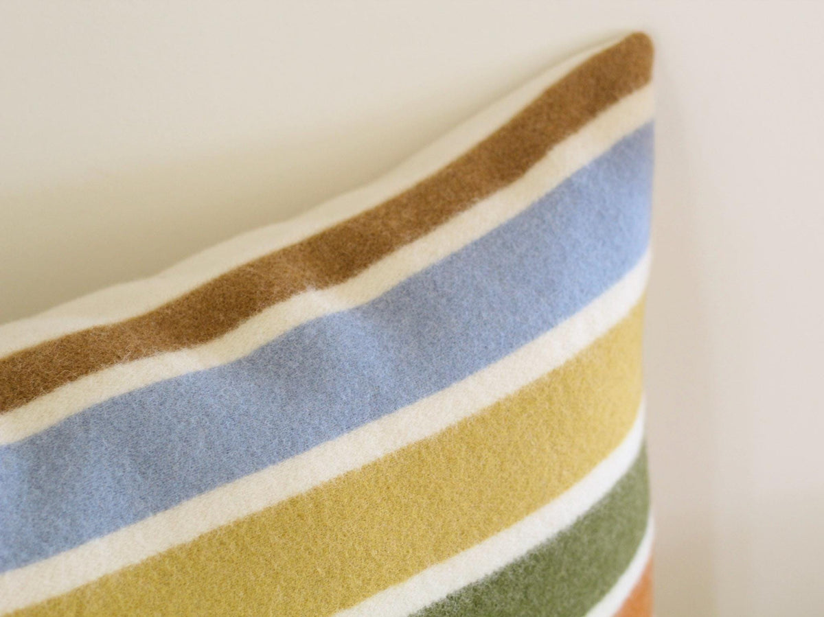 Close-up of a decorative pillow with a striped pattern. The stripes include light blue, white, yellow, green, and orange on a camel brown background. The pillow is made of a soft, textured fabric.