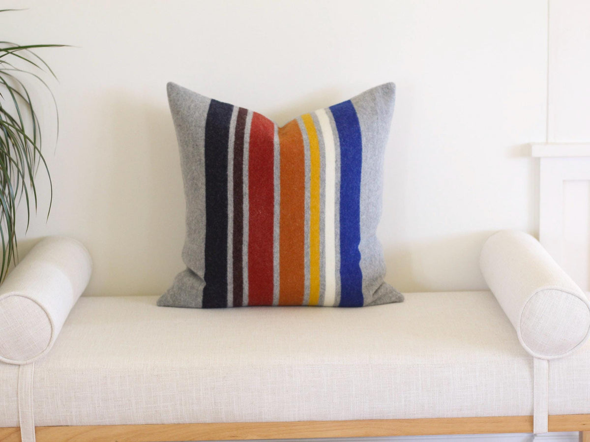 A square throw pillow featuring a vertical striped pattern in shades of gray, navy, brown, red, orange, yellow, white, and blue. The pillow has a textured, felt-like appearance and is centered on a light-colored upholstered bench.