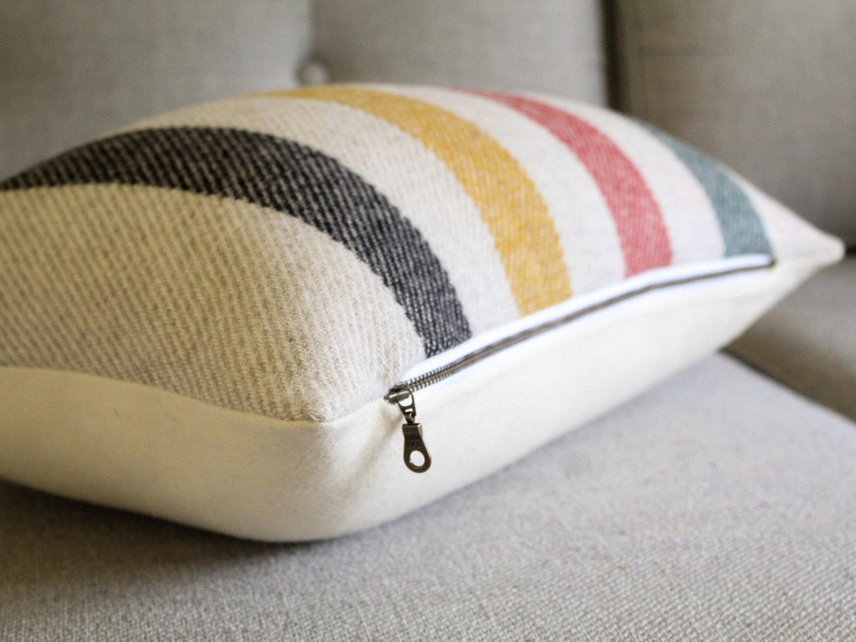 Rectangular decorative pillow with an oatmeal cream background and a striped pattern in black, yellow, red, and green. The pillow has a solid cream back and an exposed metal zipper closure and is placed on a gray sofa. The pillow is designed for home decor.