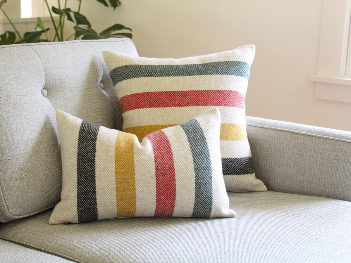 Two decorative throw pillows with a striped pattern in black, yellow, red, and green on a oatmeal and cream background. The pillows are placed on a gray sofa, creating a cozy and inviting atmosphere. The pillows add a pop of color and texture to the living space.