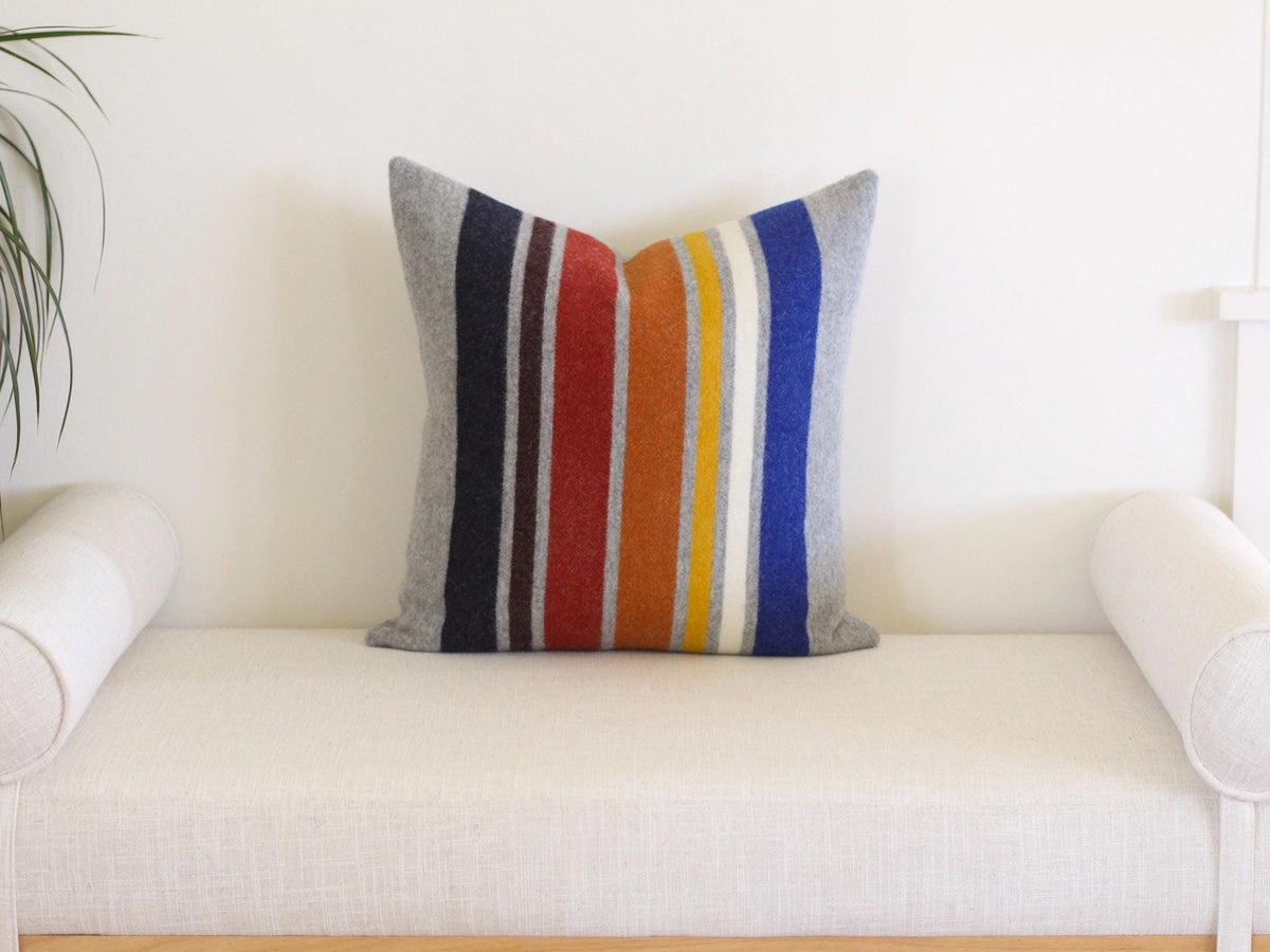 A square throw pillow featuring a vertical striped pattern in shades of gray, navy, brown, red, orange, yellow, white, and blue. The pillow has a textured, felt-like appearance and is centered on a light-colored upholstered bench.