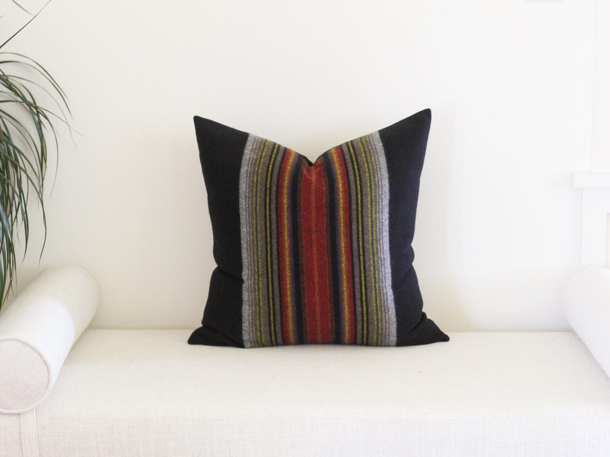 A square throw pillow featuring solid black side panels and a central vertical band with multi-colored stripes in shades of red, orange, yellow, green, and grey. The pillow has a structured shape with sharp corners and a smooth texture.